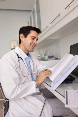 Steteskoplu beyaz önlük giyen genç iyimser erkek doktor, modern tıp ofisinde masasında oturuyor, hasta dosyalarına veya sağlık raporlarına bakarken gülümsüyor ve olumlu sonuçları inceliyor.