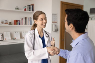 Üniformalı kadın pratisyen hekim, erkek hastayla el sıkışıyor, hastanede tıbbi bölüşmeyi başarıyla başlatıyor ya da tamamlıyor. Saygı, minnettarlık ve dostça ilişkiler gösteriyor.