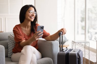 Gözlüklü genç İspanyol bir kadın seyahat başvurusunda tatil için evini arıyor, akıllı telefondan online rezervasyon hizmeti veriyor, cep telefonu kullanıyor, dişlek tebessümle başka yere bakıyor.