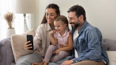 Gadget 'la güzel bir aile zamanı. Anaokulu kızı olan mutlu evli çiftler kanepede sarılır video görüşmesi için akıllı telefon kullanır birlikte şirin selfie çeker sosyal medyadan eğlenceli videolar izlersiniz.