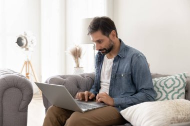 Ev işlerinden profesyonelce. Odaklanmış orta yaşlı işadamı oturma odasındaki kanepeye dizüstü bilgisayar kullanıyor online iş eğitimi alıyor internette geziniyor sohbet alışverişinde iletişim kuruyor.