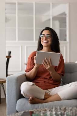 Dijital internet teknolojisinden zevk alan genç Latin kadın, evde çalışan akıllı uygulama, tablet bilgisayar kullanan, oturma odasına bakan, gülümseyen, gülen. Dikey çekim