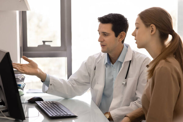 Professional medical worker explaining diagnose, treatment plan to woman patient, sit at desk, points on computer screen, provide explanation, advice or recommendation, showing new healthcare platform