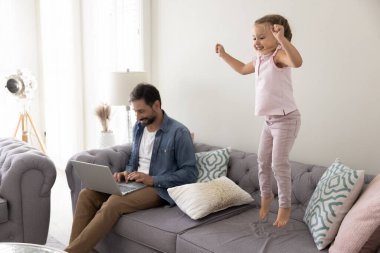 Serbest iş ve ebeveynlik. Gülümseyen orta yaşlı baba uzaktan kumandayla internetteki iş için not defteri kullanıyor. Aktif, oyuncu bir kız çocuk gibi kanepede oturuyor ve yakınlarda gülüyor.