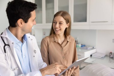 Beyaz üniformalı arkadaş canlısı erkek doktor danışma sırasında neşeli genç bayan hastaya tablet cihazıyla tıbbi bilgi veriyor. E-sağlık, teletıp platformu, modern klinikte dijital bakım