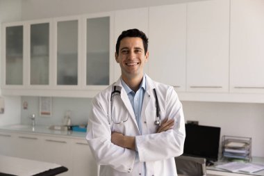 Üniformalı gülümseyen genç erkek doktorun portresi, sağlık sektöründe güven, profesyonellik ve bakım saçan kendine güvenen duruş ve dostça ifadeyle kollarını kavuşturarak ayakta duruyor.