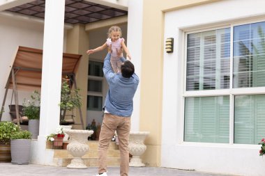 Konforlu evde aile bağı. Neşeli baba, gülen kız çocuğunu kaldırarak evin ön bahçesinde, tahterevalli ve çiçeklerle dolu güneşli verandanın yanındaki oyun saatinin tadını çıkarıyor. Boşluğu kopyala