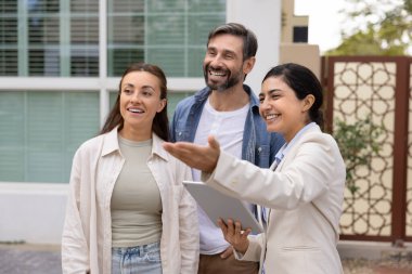 Sanal ve gerçek zamanlı ev gösterimi. Neşeli kadın emlakçı karı koca müşterileri ile bahçede dikiliyor. Ev sahiplerine ev kiracıları için dijital jest yapıyorlar.