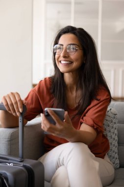 Gözlüklü genç Latin bayan turist bavullarını ve akıllı telefonlarını evde tutuyor. Daire ayırtmayı, uçak biletini, tatili, seyahati, başka yere bakmayı, gülümsemeyi düşünüyor. Dikey çekim
