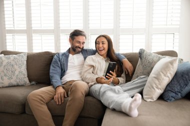Dijital eğlence ve rahatlama. Neşeli, orta yaşlı çift hafta sonu kanepesinde oturup komik mim videosu sesine ya da cep telefonundaki komik sosyal medya içeriğine gülüyor.