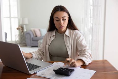 Dijital çağda finansal planlama. Meşgul genç bir kadın muhasebe işlemleriyle uğraşıyor. Rahat ev ofisinin defter hesap makinesiyle. Faturaları hesaplıyor.