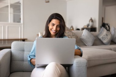 Mutlu güzel İspanyol kadın internet iletişimini evde eğlence olsun diye kullanıyor, dizüstü bilgisayarda yazıyor, gülüyor, rahat koltukta rahatlıyor, online teknolojiyle eğleniyor.