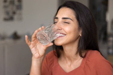 Neşeli genç İspanyol kadın kapalı gözlerle temiz suyun tadını çıkarıyor, soğuk içecekle ferahlatıcı dişlek gülümsemeyle, şeffaf camdan su içiyor, sağlıklı su ihtiyacını karşılıyor.