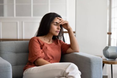 Üzgün genç Latin bir kadın kafasını kapalı gözlerle tutuyor, koltuğunda oturuyor, sorunları, sorunları, kötü haberleri düşünüyor, endişeli, endişeli, baş ağrısından muzdarip.