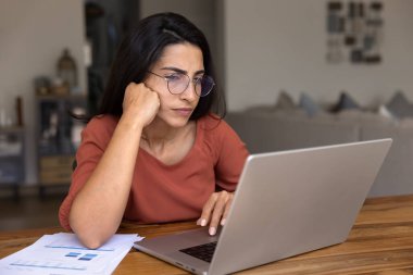 Kafası karışmış, gözlüklü İspanyol serbest çalışan, sorun çözme konusunda düşünen, dizüstü bilgisayarına yaslanan, online pazarlama stratejisini düşünen, finansal satış raporlarını hazırlayan,