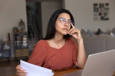 Ciddi, düşünceli, İspanyol, gözlüklü, ev bilgisayarında iş yerinde bilgisayarını düşünen, yüzüne dokunan, derin düşüncelere dalan, elinde kağıt belgeler tutan serbest bir kadın.