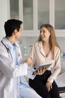 Gülümseyen kadın hasta muayene masasında oturuyor ve elinde dijital tablet tutan neşeli erkek doktorla tokalaşıyor. Modern klinikte güven, güvenilirlik ve olumlu iletişim.