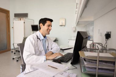 Tıp bürosundaki bilgisayarda çalışan beyaz önlük giyen gülümseyen doktor, hastalarla olumlu çevrimiçi istişareler sunuyor, iyi haberleri paylaşıyor ya da okuyor, klinikte verimli iş akışı sağlıyor.