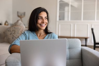 Evinde dizüstü bilgisayarda çalışan neşeli, hayalperest genç Latin serbest kadın kucağında bilgisayarla oturuyor, dişlek gülümsemesiyle başka yere bakıyor, online yaratıcı projesini düşünüyor.