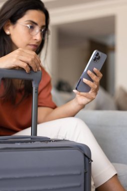 Arka planda bulanık bir şekilde otel odasında cep telefonu kullanan bir kadının bavulunun dikey görüntüsü. Ciddi genç Latin bayan turist tatil için yer ayırtıyor, uçak biletleri alıyor.
