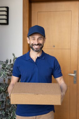 Profesyonel kurye servisi. Dikey portre mutlu teslimatçı şirket üniforması ve şapka giymiş müşteri ev kapısında poz karton kutu ve posta paketi ile gülümseyerek kameraya bakıyor