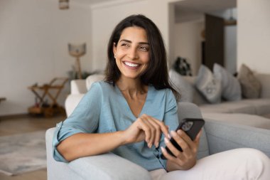 Mutlu rüya gibi Latin kadın çevrimiçi alışveriş yapmayı düşünüyor, akıllı telefon kullanıyor, evdeki koltukta dinleniyor, dişlek tebessümle başka yere bakıyor, internet servisinin keyfini çıkarıyor, modern teknoloji