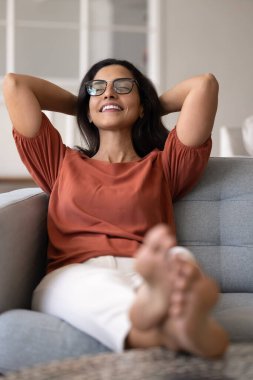 Sakin, tembel, genç, İspanyol bir kadın, rahat bir evde, rahat, yumuşak bir kanepede uzanmış, arkasını yaslamış, dişlek bir gülümseme ile dinleniyor. Dikey çekim