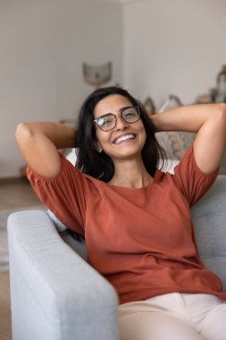Gözlüklü neşeli İspanyol kadın rahat koltukta dinleniyor, evinde dinleniyor, dişlek tebessümle kameraya bakıyor, rahatlığın, rahatlığın, serin havanın tadını çıkarıyor. Dikey portre