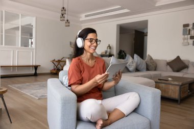 Kablosuz kulaklık takan, medyayı evinde izleyen, video konferans görüşmesinde konuşan oturma odasındaki rahat koltukta oturan, elinde aletle gülümseyen,