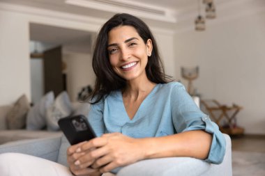 Mutlu, güzel İspanyol kadın akıllı telefonu tutuyor, rahat evdeki koltukta dinleniyor, kameraya bakıyor, gülümsüyor, portre için poz veriyor, modern internet teknolojisinden keyif alıyor, online servis kullanıyor.