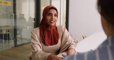 Kırmızı tesettürlü genç Müslüman profesyonel kadın toplantı masasında iş ortağıyla konuşuyor, işverenle iş görüşmesinde iş becerilerini anlatıyor, proje satıyor, gülümsüyor, konuşuyor