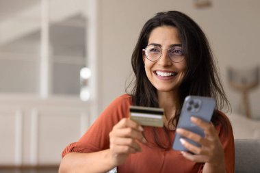 Mutlu, rüya gibi Latin banka müşterisi, gözlüklü, kredi kartı ve online ödeme başvurusunu cep telefonuyla kullanan, aleti tutan, dişlek tebessümle başka yere bakan, online alışverişin tadını çıkaran bir kadın.