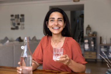 Mutlu, güzel, genç, İspanyol bir kadın elinde bir şişe maden suyu tutuyor, camdan soğuk içecek içiyor, mükemmel dişli bir gülümsemeyle fotoğraf için kameraya bakıyor, sağlıkla ilgileniyor, detoks, sıvı kaybı...