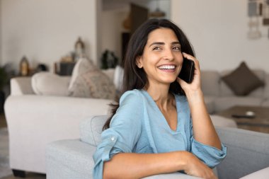 Neşeli genç Latin akıllı telefon kullanıcısı kadın cep telefonuyla konuşmaktan hoşlanıyor, cep telefonuyla konuşuyor, rahat koltukta oturuyor, gülümsüyor, gülüyor, başka tarafa bakıyor.