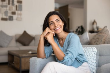 Neşeli, güzel, genç, İspanyol bir kadın rahat bir evde rahat bir koltukta oturuyor, başını kaldırıyor, başka tarafa bakıyor, düşünüyor, dişlek bir gülümsemeyle hayal ediyor, ev keyfi yapıyor.