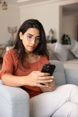 Şık gözlüklü genç Latin akıllı telefon kullanıcısı kadın evde cep telefonuyla yazı yazıyor, mesajlaşıyor, internetten sohbet ediyor, internetten alışveriş yapıyor, sipariş veriyor, web servisini kullanıyor. Dikey çekim