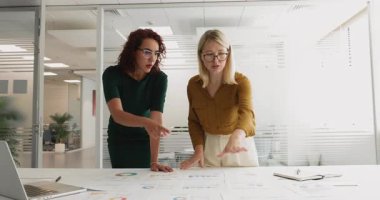 İki meşgul genç kadın proje yöneticisi toplantı masasında kağıt pazarlama raporlarını inceliyor, anlaşma yapıyor, işi tamamlıyor, yumruk tokuşturma dostluğu hareketi yapıyor, gülümsüyor.