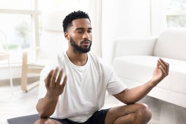 Barışçıl Afrikalı adam, rahat oturma odasında hasıraltı edilmiş, iç huzuru, odaklanmayı ve zihinsel berraklığı artırarak, farkındalık pratikleriyle denge ve sakinliği bularak, iç içe geçmiş meditasyon giysileri giyiyor.