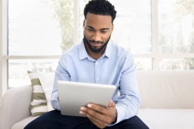 Genç 30 'lu yaşlardaki odaklı Afrikalı adam modern dijital tablet cihazını kullanarak çevrimiçi çalışıyor, müşteriye e-posta atarak, uzaktan çözerek, hobi ve eğlence için yeni bir platformun keyfini çıkararak koltukta oturuyor.