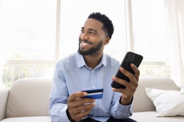 Mutlu Afrikalı adam evde online alışverişi seviyor, akıllı telefon ve banka kartıyla kanepeye oturuyor, neşeli bir ifadeyle yan yana bakıyor, alışveriş yapıyor, pazar yeri uygulamasını kullanarak hediyeler alıyor.