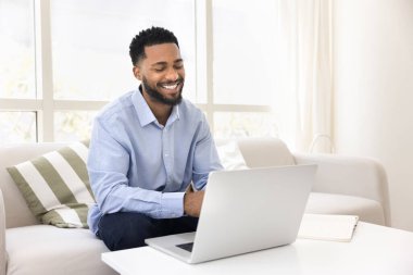 Kanepede dizüstü bilgisayarla oturan Afrikalı bir adam, online çalışıyor, video görüşmesine katılıyor, kişisel ya da işle ilgili meselelerle sanal bir toplantı yapıyor, uzaktan kumanda işini verimli bir şekilde yapıyor. Evden çalışma, iletişim.