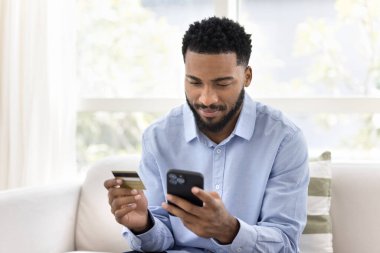 Genç Afrikalı adam internetten mal alıyor, banka kartı ve cep telefonu tutuyor, internetten hediyeler alıyor, dijital işlem yapıyor, mobil uygulama üzerinden para gönderiyor, faturaları ödüyor, rezervasyon hizmetleri ayarlıyor.