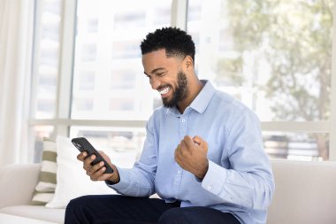 Akıllı telefon ekranına bakarken gülümseyen adam, kutlama jestinde yumruğunu sıkar, olumlu haberler alır, projenin onayını alır, maddi kazanç, fırsat, kişisel başarı veya piyango kazanır.