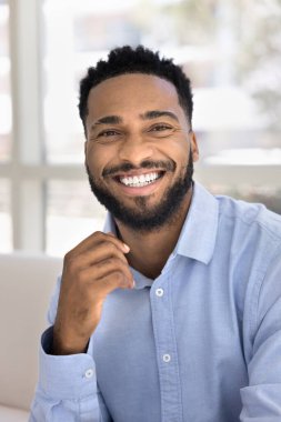 Dikey çekim, güzel görünümlü mutlu Afrikalı sakallı adam, beyaz dişli gülümseme, iyimser ruh hali, kameraya bak kanepeye otur, tasasız hafta sonunun tadını çıkar, evde vakit geçir. Ev resmi