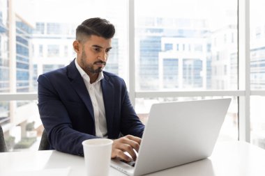 Ciddi Hintli genç yönetici bir fincan kahvede dizüstü bilgisayarda yazıyor, büyük ofis penceresinde bilgisayarın başında çalışıyor, iş iletişimi için internet teknolojisini kullanıyor. Dikey çekim