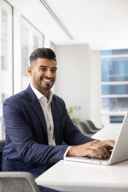 Neşeli, yakışıklı, şık, genç Hintli işadamı ofiste dizüstü bilgisayarla yazı yazıyor, göz kamaştırıcı bir gülümsemeyle, gülüyor, çevrimiçi ürün üzerinde çalışıyor, yaratıcı fikir ve çözüm üzerinde düşünüyor. Dikey çekim