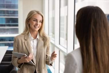 Pozitif dost canlısı sarışın profesyonel kadın ofiste bayan meslektaşıyla konuşuyor, toplantı odasında duruyor, iş ortağı, şirket müşterisiyle konuşuyor, gülümsüyor, tableti tutuyor.