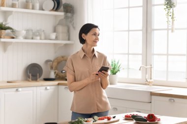 Yemek ve akıllı telefon. Gülümseyen olgun kadın, rahat bir mutfak tezgahında duruyor. Telefonu tut. Sağlıklı mutfak ve yemek fotoğrafçılığı hobilerini birleştirip sosyal medyadan yeni sebze tariflerini kontrol et.