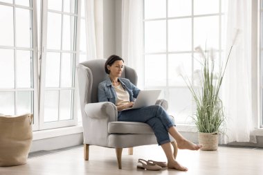 Evden çevrimiçi olarak rahat. Tam boy portre rahatlamış yaşlı kadın dinlenme odasında rahat koltukta rahat modern oturma odasında sıcak zemin işi ile birlikte uzaktaki web sitesinden not defteri pc kullanarak blog yazmayı öğrenin