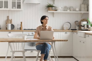 Serbest problem çözme. Düşünceli, orta yaşlı, serbest çalışan kadın mutfaktaki ofisteki defter üzerinde çalışmaktan vazgeçti. Mesafe planına bak. Proje stratejisi bütçesini analiz et.